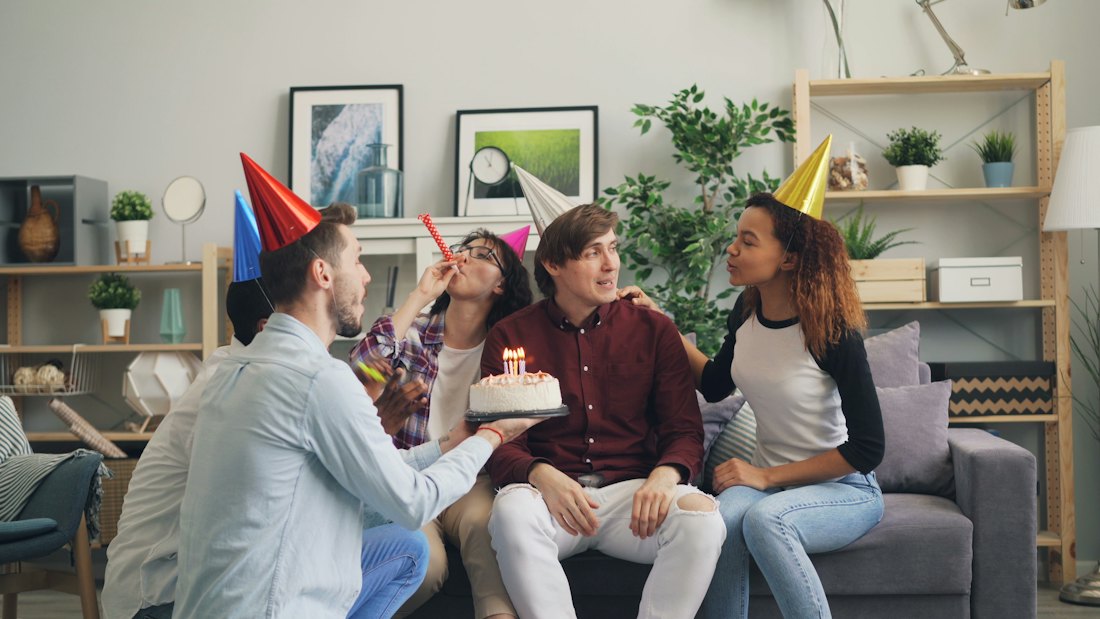 Friends blowing party horns around a birthday cake on a sofa